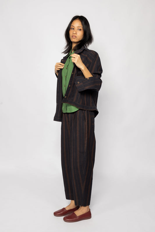 Plains Design - Brick Shirt. Oversized striped shirt in black and brick colours with two square flap pockets on the chest. The shirt has contrasting stripes in a different direction on the hem. Paired with ankle length pants in the same fabric and brown leather shoes.