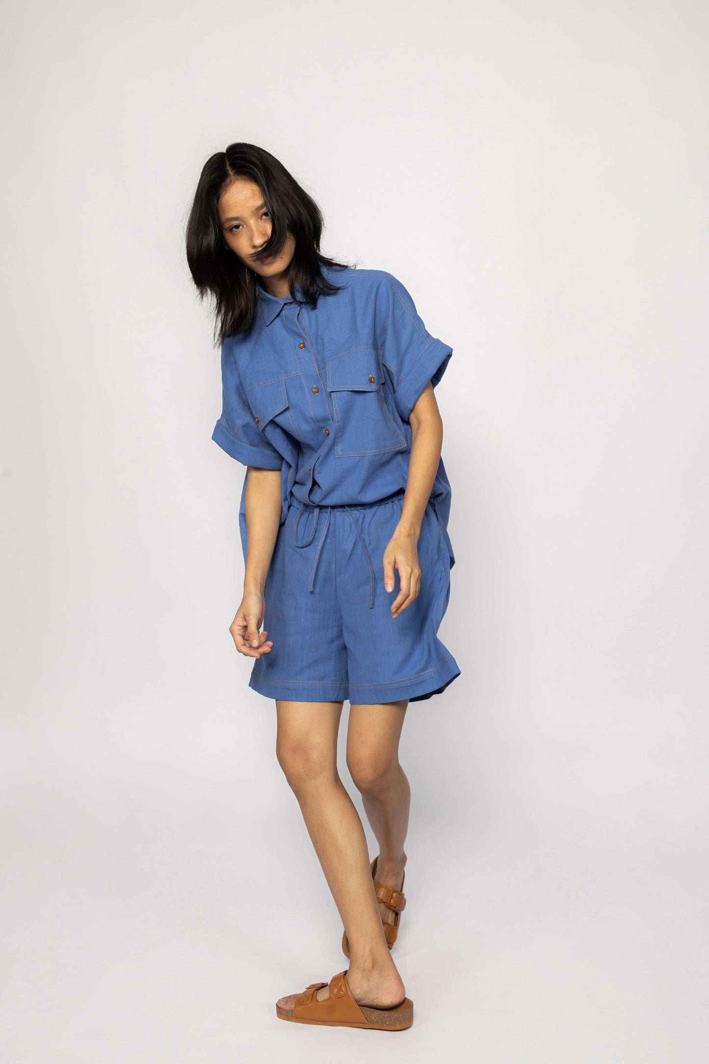 Plains Design - Trucker shirt. A person standing wearing an oversized blue shirt with orange stitching, wooden buttons, short sleeves, and two chest pockets. They are also wearing blue shorts and brown sandals.
