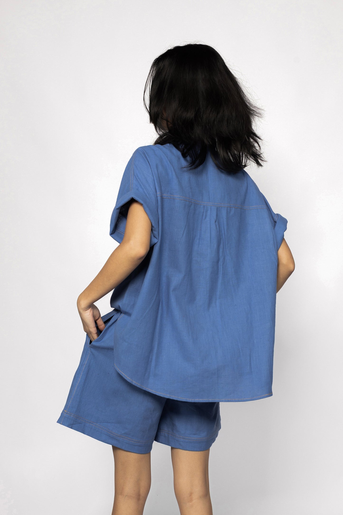 Plains Design - Trucker shirt. Back view of a person standing wearing an oversized blue shirt with orange stitching, and blue shorts. Sustainable handmade cotton.
