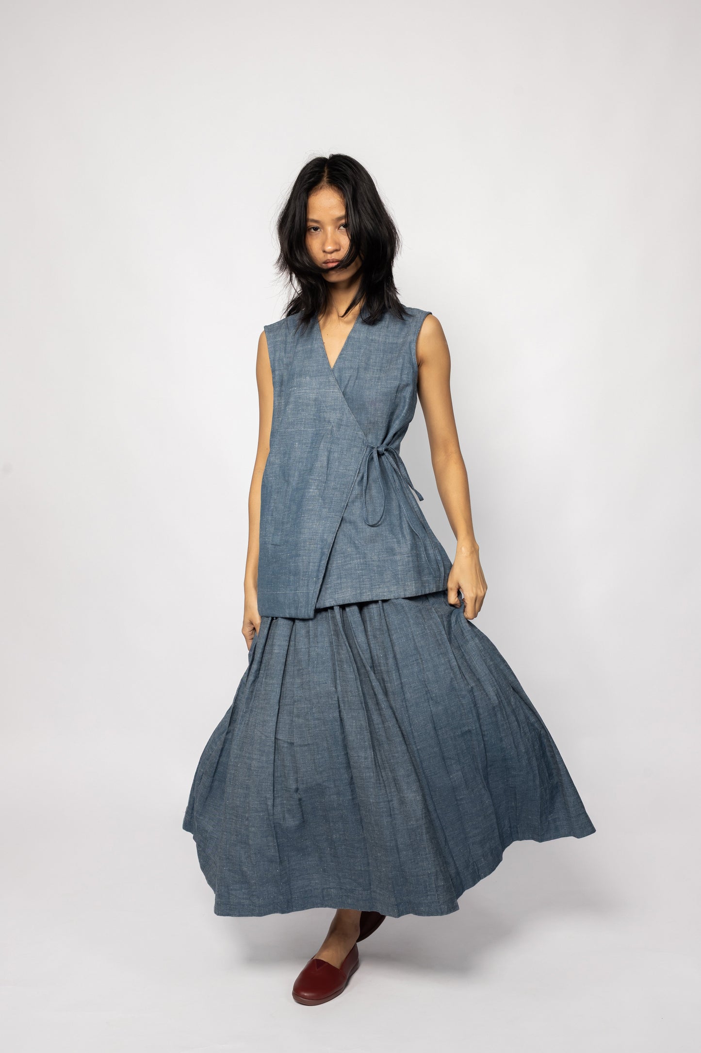 Plains Design - Lake Denim Skirt. Midi-length denim skirt with a pleated design. She is also wearing a matching denim sleeveless top. The denim is handmade using sustainable techniques.