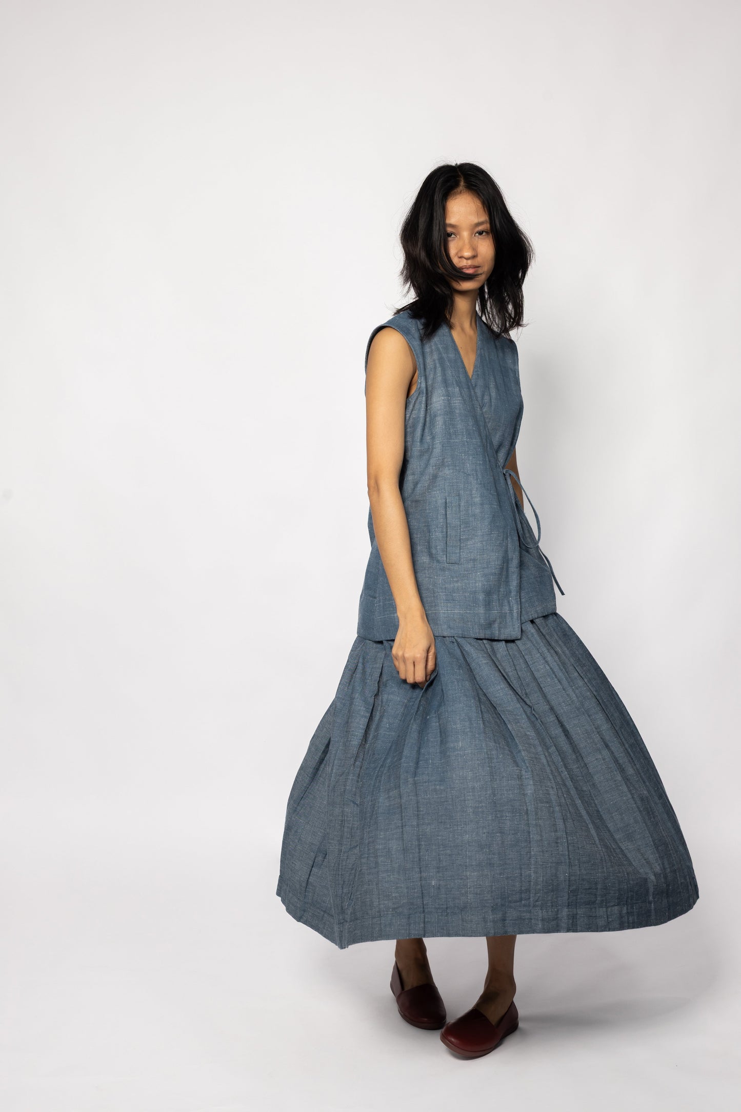 Plains Design - Lake Denim Skirt. Midi-length denim skirt with a pleated design. Paired with a matching denim sleeveless top and brown leather shoes. The denim is handmade in cotton twill.