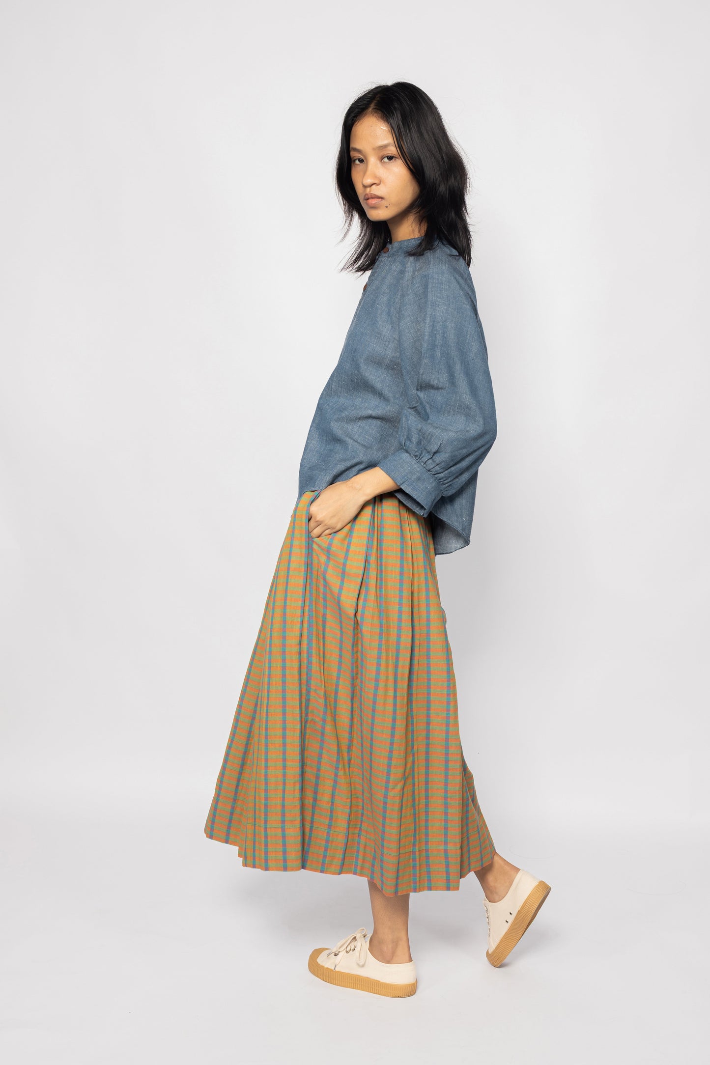 Plains Design - Twin skirt in Orange. Side profile of midi skirt with orange, blue and yellow checks in a relaxed fit and side pockets. Paired with a denim top. Ethically handmade with handspun cotton in India.