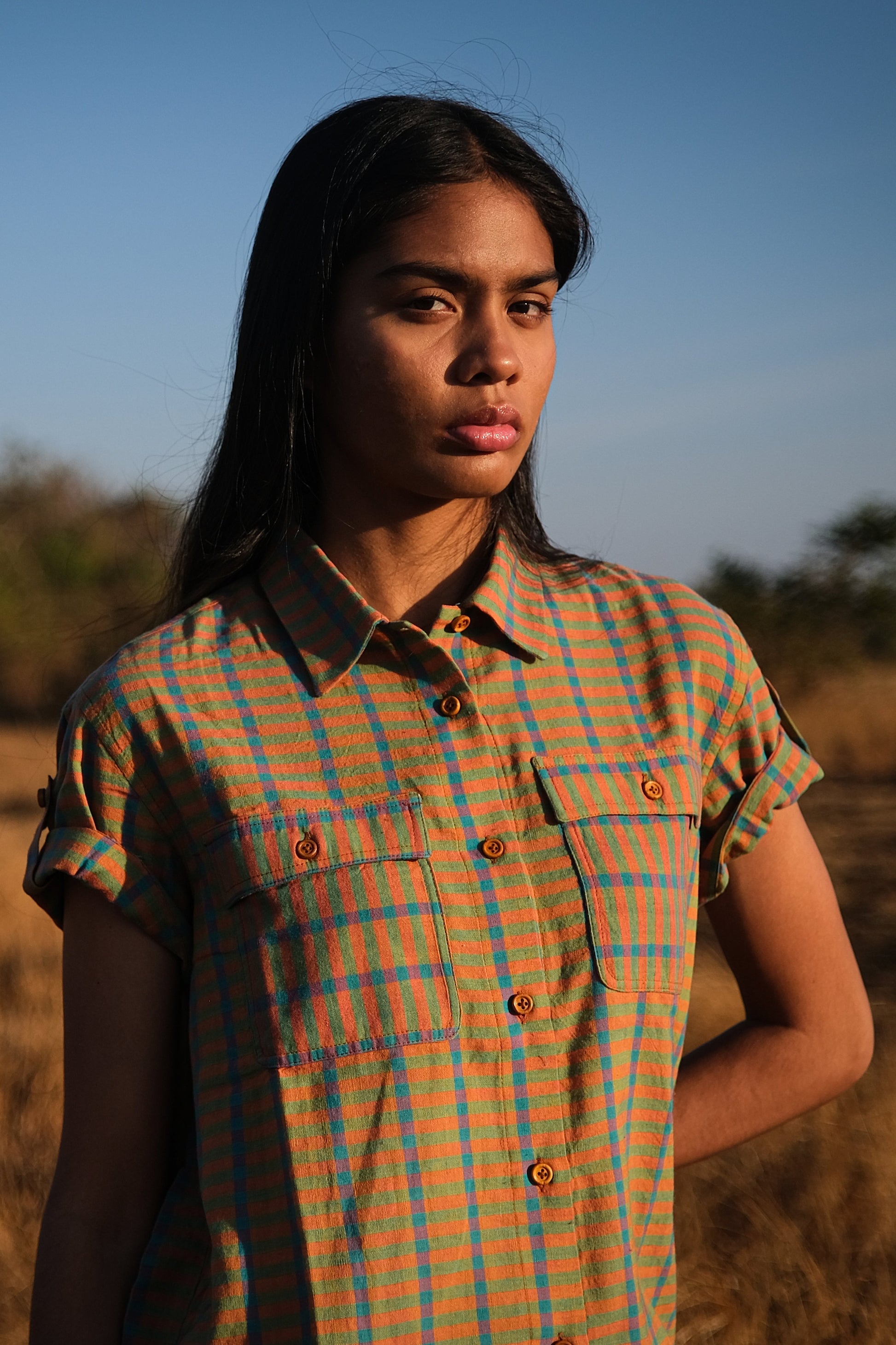 Plains Design - Twin shirt in orange. Front closeup of orange and blue checkered shirt with short folded sleeves, two chest pockets, collar, and wooden buttons in a straight relaxed fit. Ethically handmade cotton.
